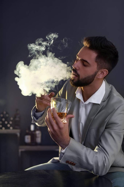 Man with glass of whiskey and cigar sitting at table indoors - Foto, afbeelding