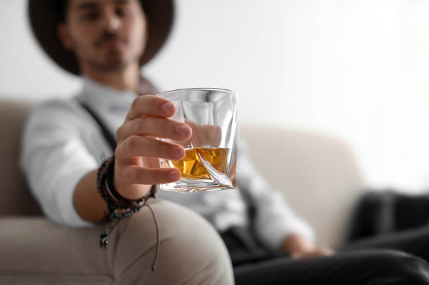 Young man with glass of whiskey sitting on sofa at home. Space for text - Фото, зображення