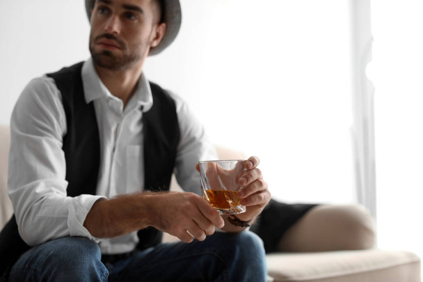 Young man with glass of whiskey sitting on sofa at home - Fotografie, Obrázek