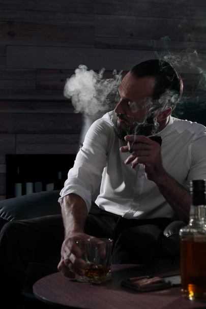 Man met glas whiskey en sigaar zittend aan tafel binnenshuis - Foto, afbeelding