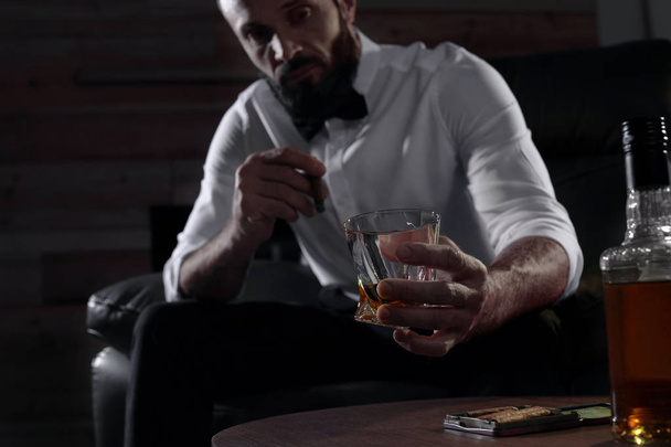 Hombre con vaso de whisky y cigarro sentado en la mesa en el interior
 - Foto, Imagen