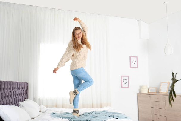 Attractive smiling young woman in cozy warm sweater jumping on bed at home - Foto, Imagem