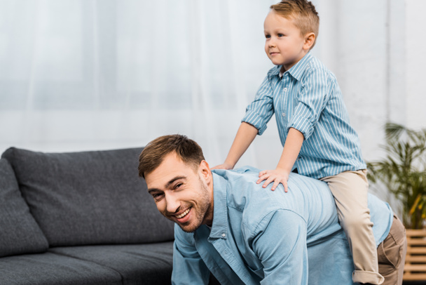 father standing on all fours and looking at camera while cute boy sitting on back in apartment - Photo, Image