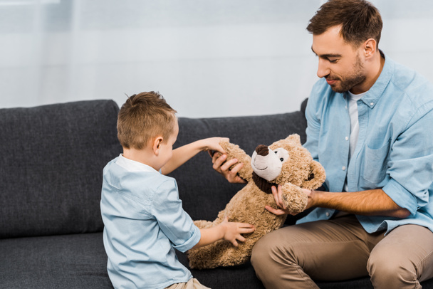 smiling father and son sitting on sofa and holding teddy bear in living room - Φωτογραφία, εικόνα