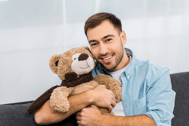 pohledný muž s úsměvem, objímá medvídka a při pohledu na fotoaparát v bytě - Fotografie, Obrázek