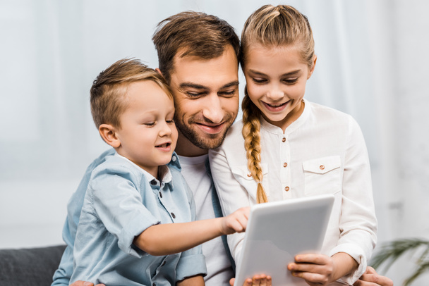 smiling father embracing cute girl holding digital tablet and adorable boy pointing with finger at screen in living room - Foto, Imagem