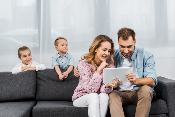 cute siblings looking at parents sitting on sofa and using digital tablet in living room - Foto, immagini