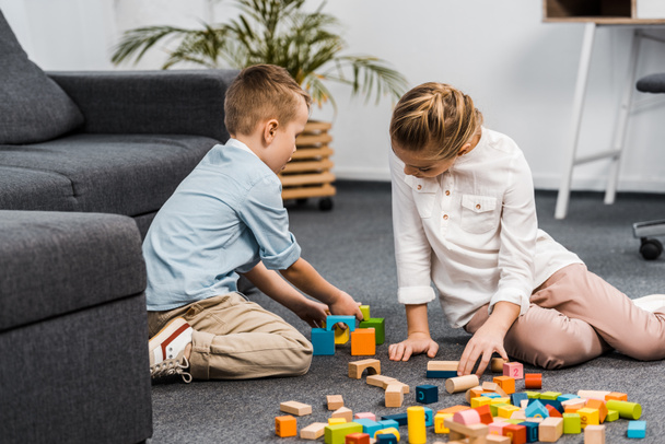 cute girl and boy sitting on floor and playing with multicolored wooden blocks in apartment - Photo, Image