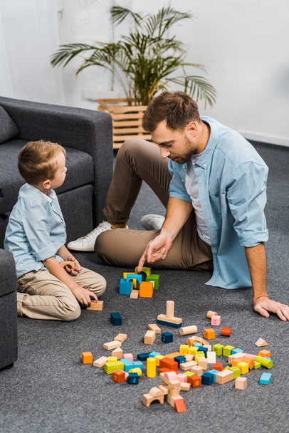 handsome father and cute son playing with multicolored wooden blocks on floor in living room - Фото, изображение