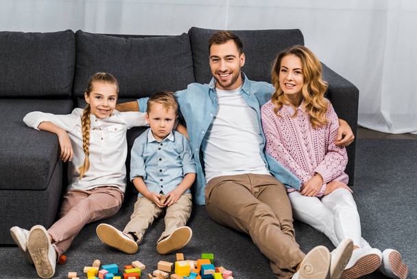 smiling father, mother, daughter and upset son sitting on floor near sofa in apartment - Foto, afbeelding