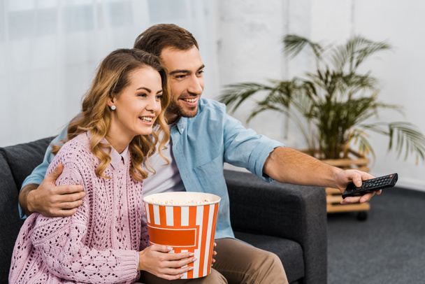 smiling man sitting on sofa, holding remote controller and embracing wife with striped popcorn bucket - Fotografie, Obrázek