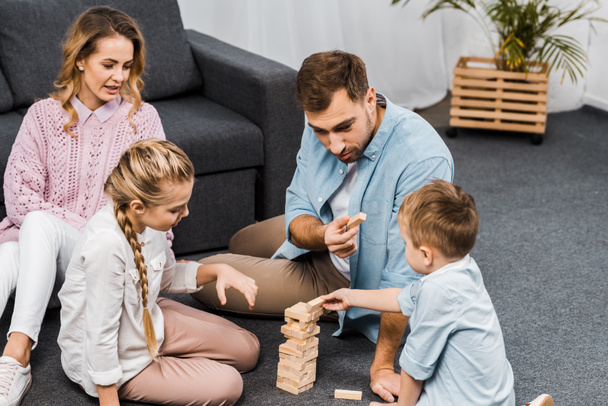 two parents playing blocks wood tower game with children on floor in apartment - Photo, Image