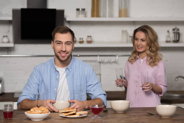 улыбающийся мужчина сидит за столом и смотрит в камеру с красивой женой, держащей очки на заднем плане
 - Фото, изображение
