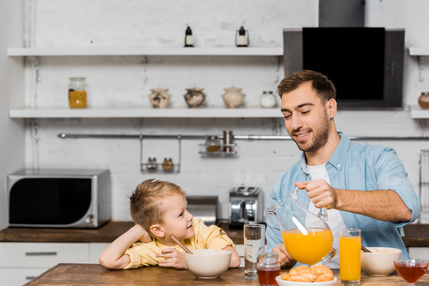 cute boy looking at smiling father pouring orange juice in glass on kitchen table - Foto, Bild