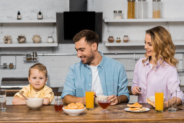 улыбающиеся родители смотрят на симпатичного мальчика, сидящего за кухонным столом
 - Фото, изображение