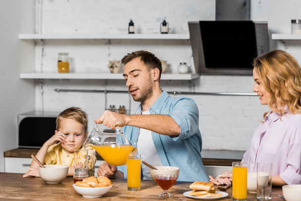 mooie vrouw en schattige jongen kijken naar knappe man gieten sinaasappelsap in glas in de keuken - Foto, afbeelding