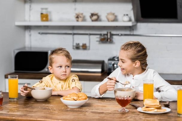 smiling girl holding spoon with porridge and looking at upset little brother at table - Foto, Imagen