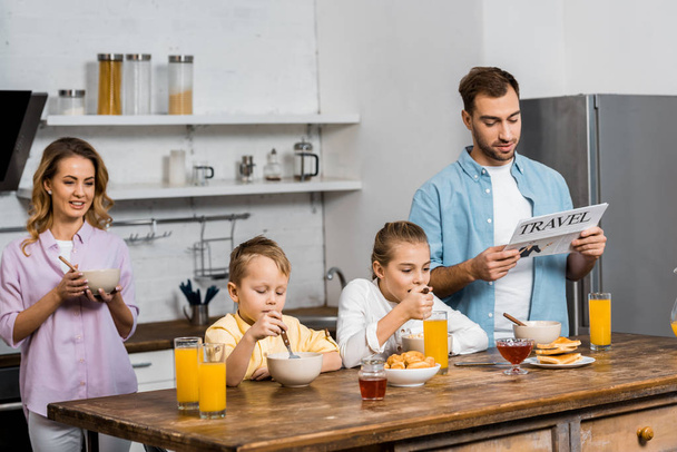 son and daughter eating oatmeal while father reading travel newspaper in kitchen - Fotó, kép