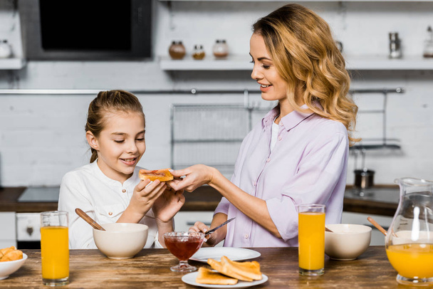 Lachende vrouw toast met jam geven met schattige dochter aan tafel in de keuken - Foto, afbeelding