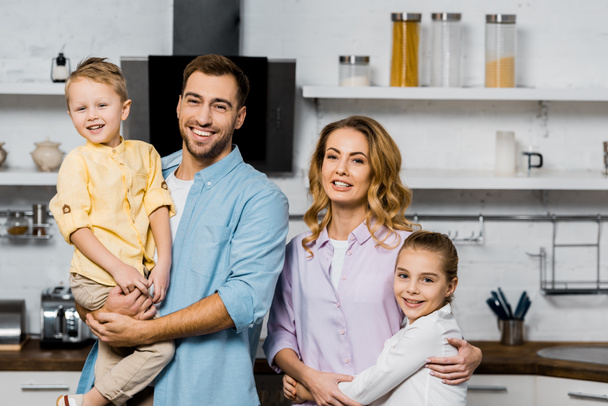 handsome man holding son and pretty mother embracing cute daughter in kitchen - Fotografie, Obrázek