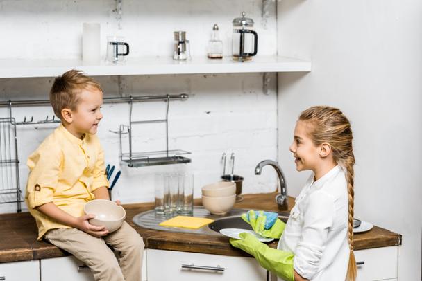 cute girl in rubber gloves washing dishes and looking at smiling boy sitting on table and holding bowl in kitchen - Фото, изображение