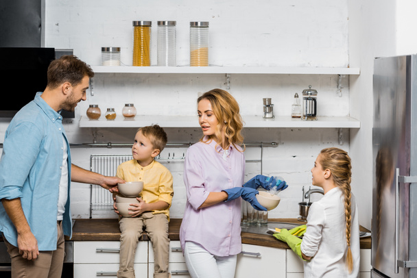 matka a dcera v gumové rukavice na mytí nádobí, zatímco otec a syn drží misky v kuchyni - Fotografie, Obrázek