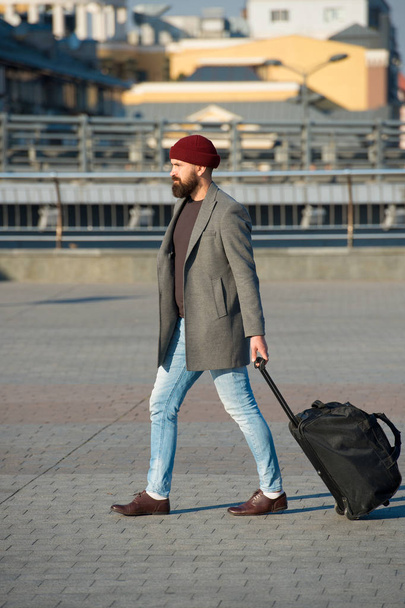 Adjust living in new city. Man bearded hipster travel with luggage bag on wheels. Traveler with suitcase arrive airport railway station urban background. Hipster ready enjoy travel. Carry travel bag - Fotoğraf, Görsel