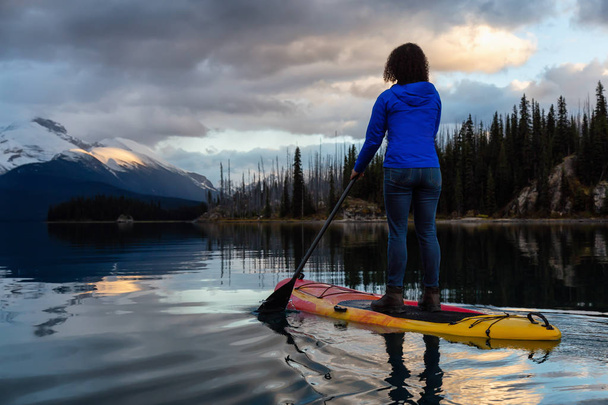 Girl Paddle Boarding in a peaceful and calm glacier lake during a vibrant cloudy sunset. Taken in Maligne Lake, Jasper National Park, Alberta, Canada. - Foto, immagini