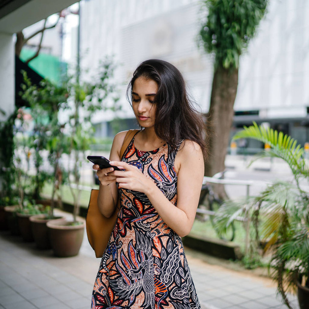 young Indian Asian woman standing and using her smartphone on an urban street in a city in Asia during the day - 写真・画像