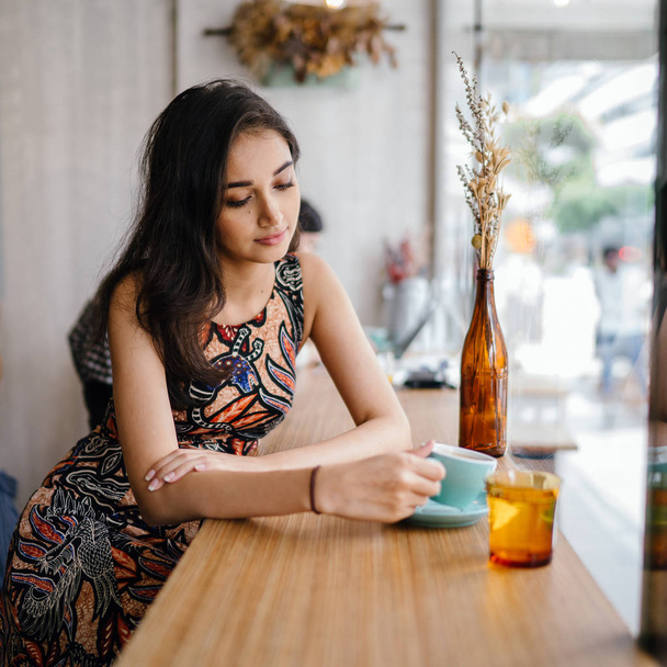 Young and beautiful Indian Asian Singaporean woman in a print dress sits in a cafe as she enjoys the weekend in Asia. - 写真・画像