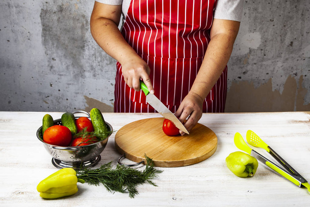 Vrouw in een rood schort is de groenten snijden voor een salade op een tafel in de keuken. Koken. - Foto, afbeelding