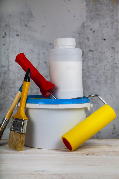 Tinta lata, rolo e escova sobre a mesa. Itens para renovação de casa ou escritório contra uma parede cinza
. - Foto, Imagem