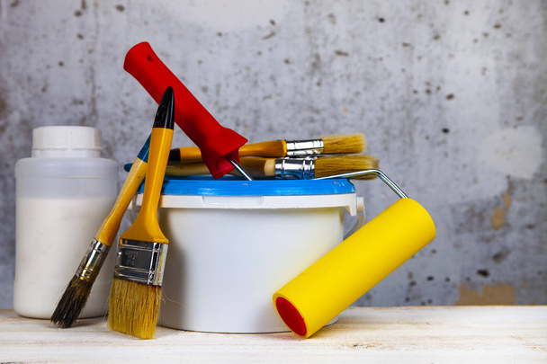 Farbdose, Rolle und Pinsel auf dem Tisch. Gegenstände für die Renovierung zu Hause oder im Büro vor einer grauen Wand. - Foto, Bild