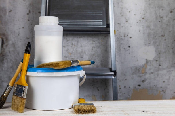 Tinta lata, rolo e escova sobre a mesa. Itens para renovação de casa ou escritório contra uma parede cinza e escada
. - Foto, Imagem