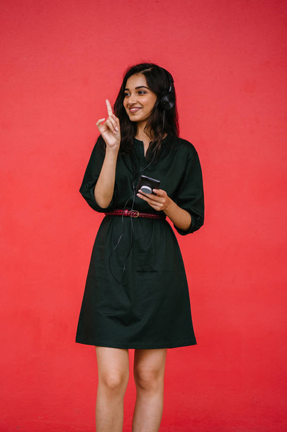 Young, attractive and beautiful Indian Asian millennial girl dancing as she listens to music on her headphones. She is streaming music on her smartphone - Foto, imagen