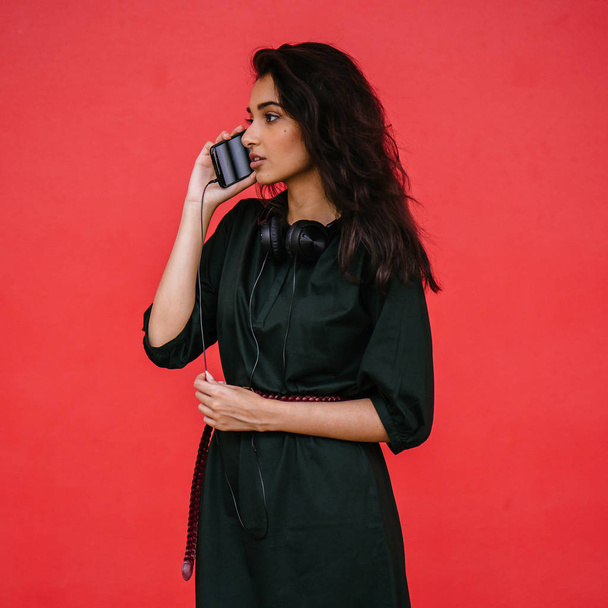 Studio portrait of a beautiful, young and attractive Indian Asian woman in a green dress and red belt against a red background. She is using her smartphone. - Φωτογραφία, εικόνα