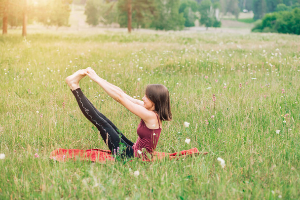 Beautiful young brunette woman in Burgundy top and black leggings doing yoga in nature - Photo, image