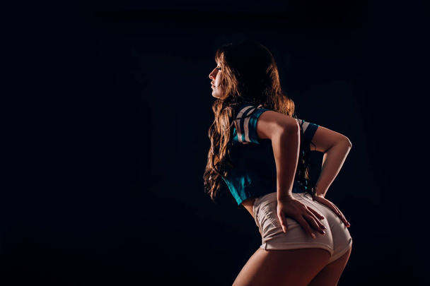Sideways portrait of sexual beautiful brunette woman in blue t-shirt, short white shorts on black background with copy space in darkness. Tanned fitness and dancer girl holds her booty with her hands - Photo, Image