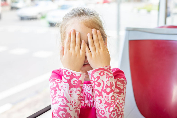 Retrato de menina de quatro anos, com camisa rosa, escondendo os olhos com as mãos e os dedos, espreitando ou brincando de esconder e procurar, sorrindo com expressão feliz no ônibus
 - Foto, Imagem