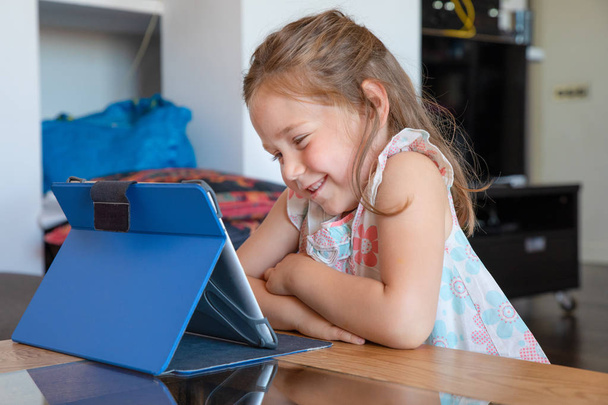 quatro anos de idade loira bonito menina sentado em interior, na sala de estar de casa, assistindo tablet digital na mesa com expressão rosto sorridente
 - Foto, Imagem