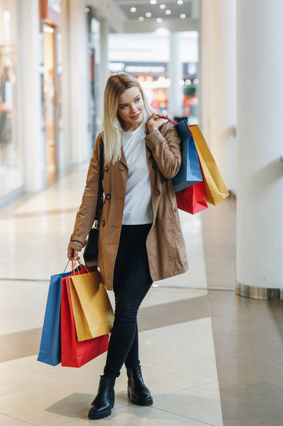 Young blond girl in brown coat whit red, blue, yellow shopping bags stay in big city shopping mall and look to store with sale. Concept: shopping, sales, Consumerism, lifestyle - Photo, Image