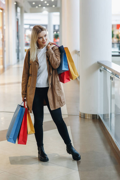 Young blond girl in brown coat whit red, blue, yellow shopping bags stay in big city shopping mall and look to store with sale. Concept: shopping, sales, Consumerism, lifestyle - 写真・画像