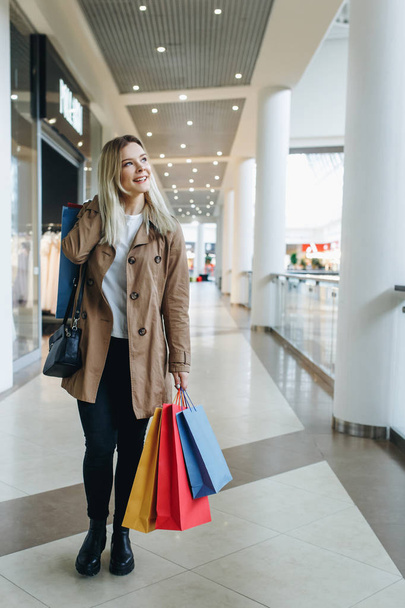 happy girl smile and walking in the big city MALL whit multicolored shopping bags. Young blond happy girl smile in the big store with shopping packages. Shopping, sales, Consumerism and lifestyle concept - 写真・画像