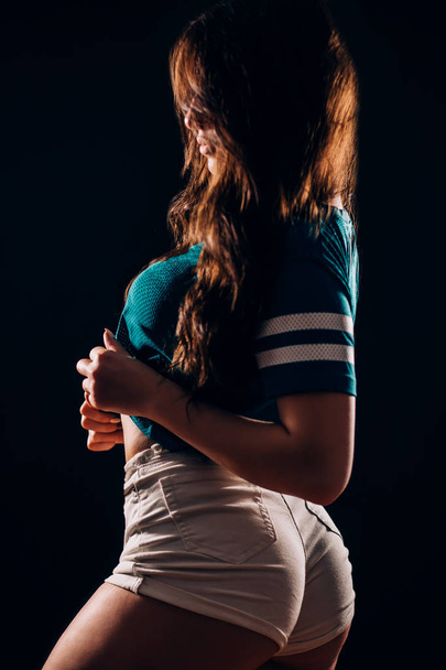 Sideways sexy portrait of young hot brunette woman in short white shorts and t-shirt in dark on black background, silhouette fitness and dancer girl - Φωτογραφία, εικόνα