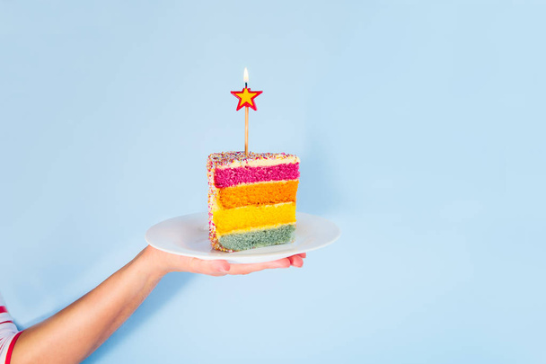Female hand holding white plate with slice of Rainbow cake with birning candle in the shape of star isolated on blue background. Happy bithday, party concept. Square card. Selective focus. Copy space - Фото, зображення