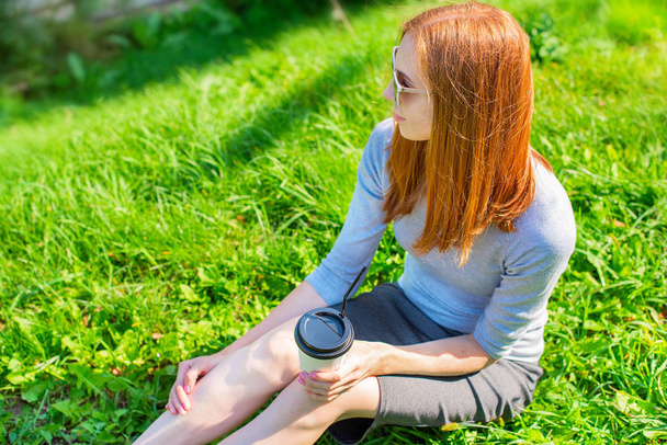 Beautiful red-haired girl sitting on the grass and drinking coffee from a paper Cup. Lifestyle Breakfast in the Park - Photo, Image