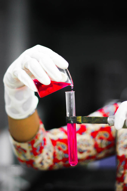 verter un líquido rojo de un vaso de precipitados a un tubo de ensayo con una mano enguantada en un laboratorio de química
 - Foto, Imagen