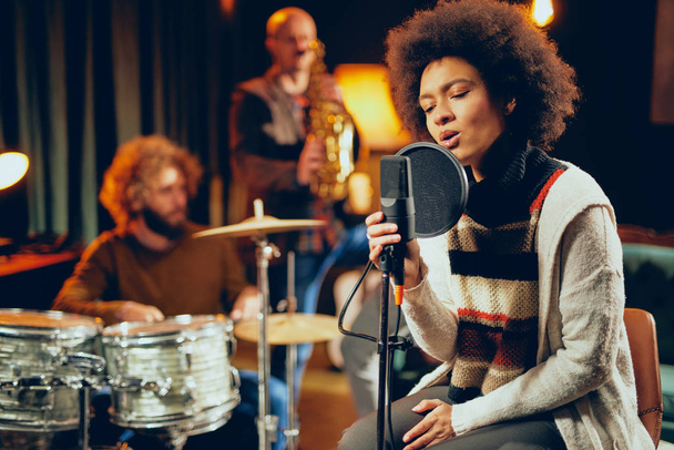 Mixed race woman singing. In background band playing instruments. Home studio interior. - Photo, Image