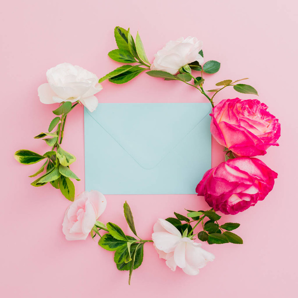 Floral frame met rozen bloemen en blauwe envelop op roze achtergrond. Plat lag, top uitzicht. - Foto, afbeelding