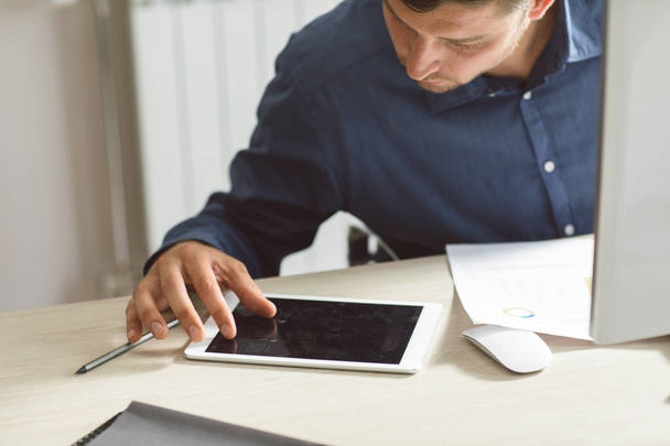 Jovem empresário usando um tablet enquanto trabalhava em um computador desktop no moderno espaço de escritório
. - Foto, Imagem